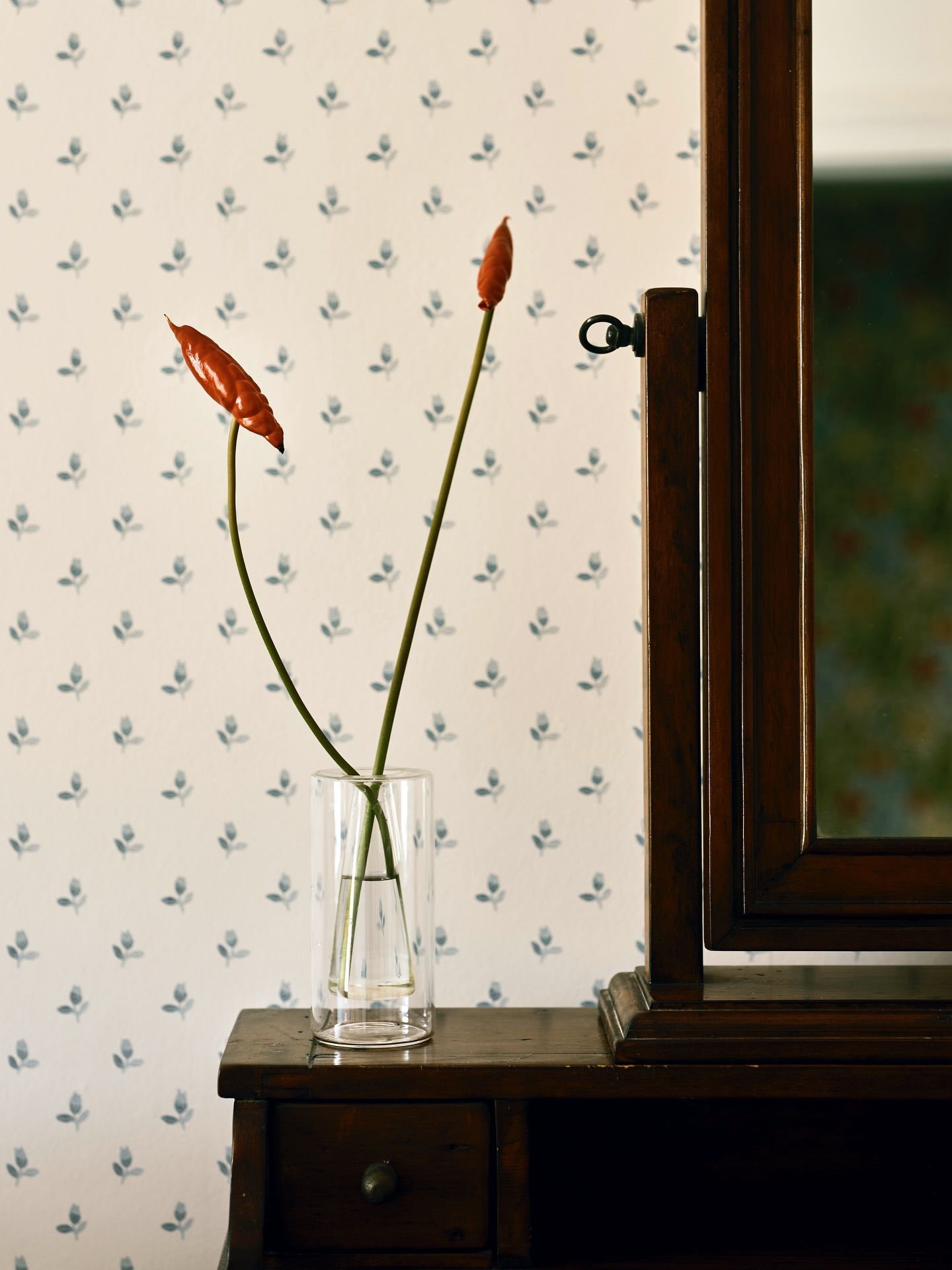 Dark timber antique dressing table with a swing mirror, styled with two sculptural red stems in a slim glass vase against sprigged wallpaper