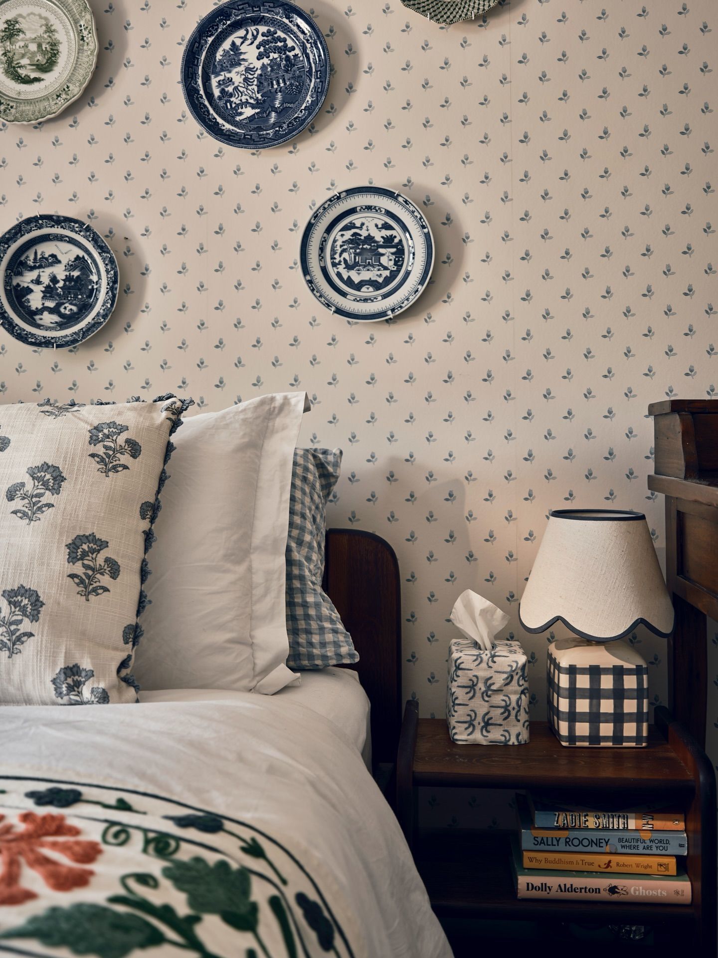 Cosy blue bedroom with antique blue-and-white plates hung on sprigged wallpaper, a gingham cushion and a scalloped lampshade on the bedside table
