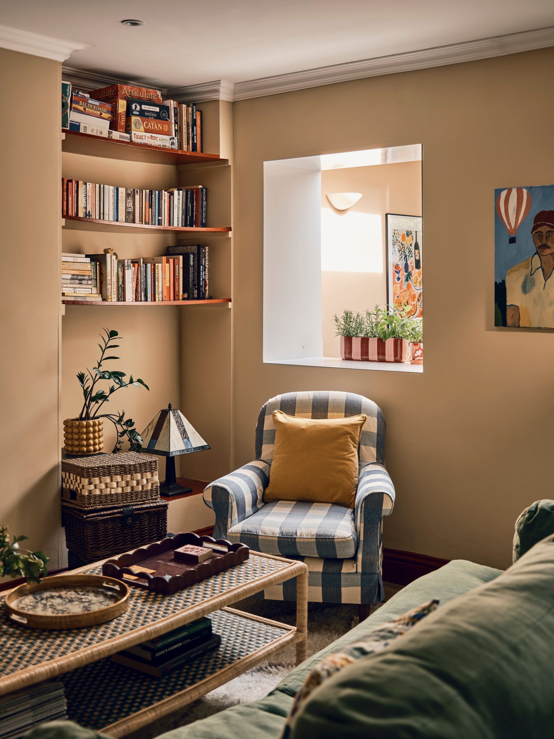Cosy snug with a blue gingham armchair, mustard velvet cushion, rattan coffee table and alcove bookshelves